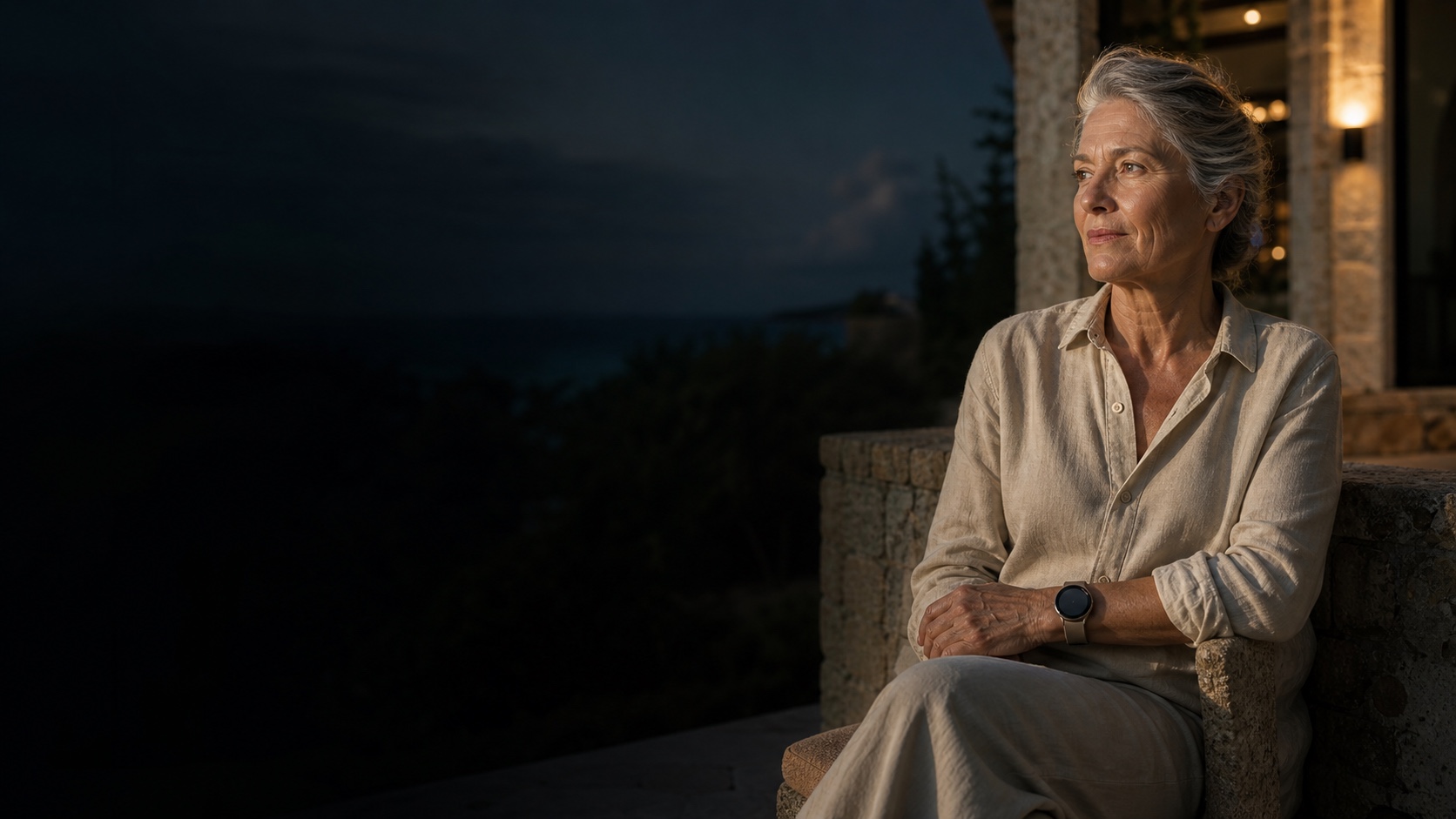 Eine selbstständige ältere Frau sitzt ruhig am Abend auf einer mediterranen Terrasse und trägt eine neutrale Smartwatch.
